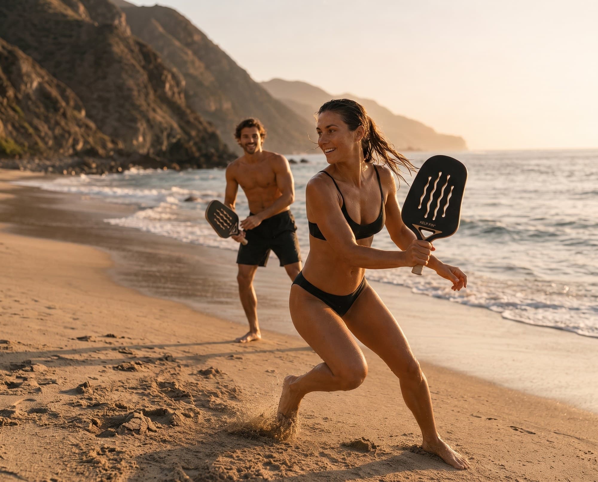 Two people playing with KELP Fins on the sand at golden hour, the coastline behind them
